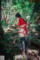 A woman in a red shirt and black stockings standing on a rock in the woods.